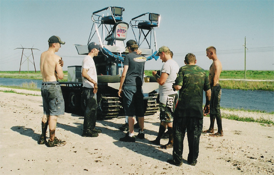 Group of muddy men around a blue tracked swamp buggy on a canal levee