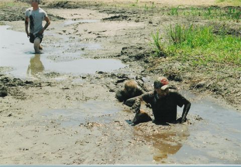 1992 Ford Bronco - Two people sit waist-deep in a brown mud hole, covered head to toe in mud, while a third person in a gray shirt wades through shallow water…