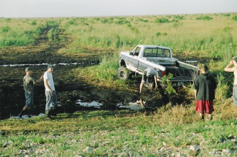 1992 Ford Bronco - A lifted grey Ford pickup on oversized mud tires sits bogged down at the edge of a black mud pit in an open grassy field, its front end ang…