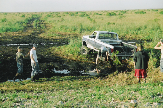 Lifted grey Ford pickup stuck in a mud pit with people gathered around it