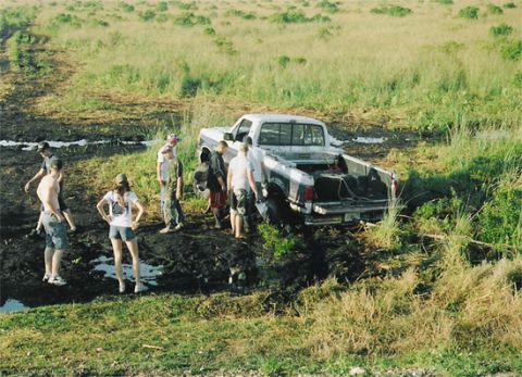 1992 Ford Bronco - Elevated view of a white extended-cab pickup bogged down to its axles in a muddy rut cut through a green grassy field, with about eight peo…