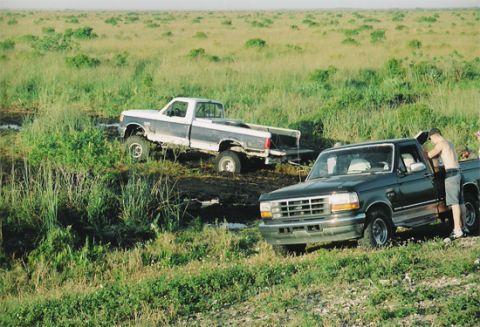 1992 Ford Bronco - Two Ford pickup trucks are stuck in a muddy, grassy field, with one truck partially submerged.