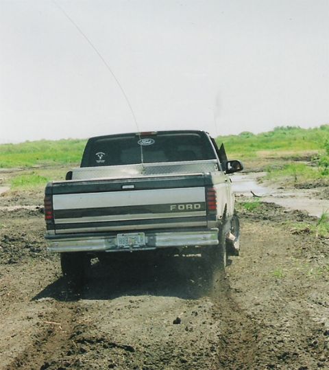 1992 Ford Bronco - Rear view of a lifted two-tone Ford pickup with a diamond-plate toolbox and a tall whip antenna, driving away on a rutted dirt and mud trac…