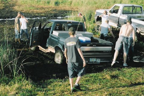 1992 Ford Bronco - A dark green extended-cab pickup with an aluminum bed toolbox and cooler is stuck in a muddy field, with a group of people gathered around…