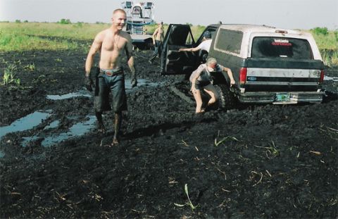1992 Ford Bronco - Scanned photo of a mud-caked Ford Bronco stuck to its axles in a black mud bog, driver's door open and a sticker on the rear window, while…