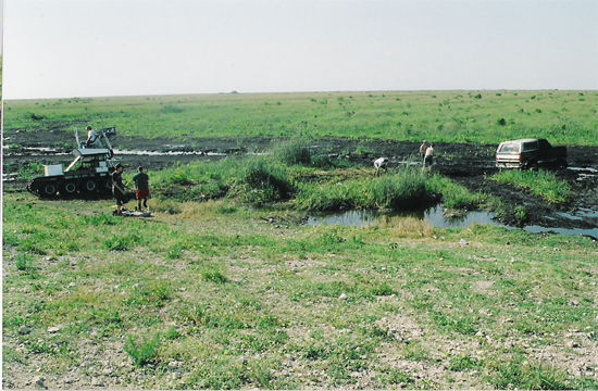 Dark SUV stuck in mud, side view, with a tracked vehicle and people nearby