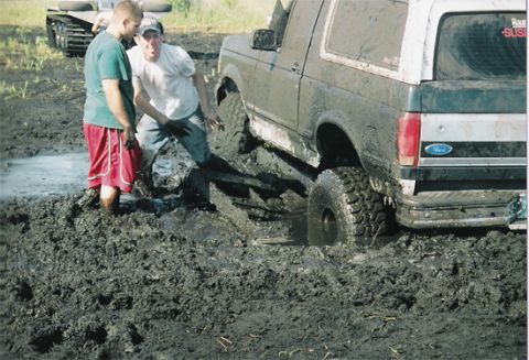 1992 Ford Bronco - Two-tone full-size Ford Bronco buried axle-deep in a churned mud pit at a mud-bogging event, its rear tailgate carrying a Ford oval and 'SU…