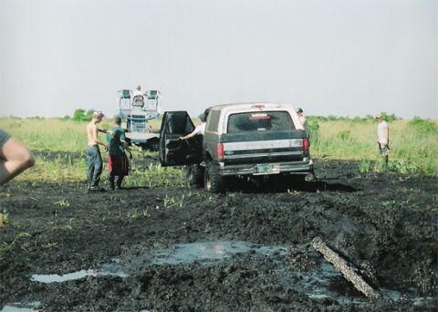 1992 Ford Bronco - Rear three-quarter view of a two-tone gray and white Ford Bronco with its driver's door open, sitting in a churned black mud bog with a red…