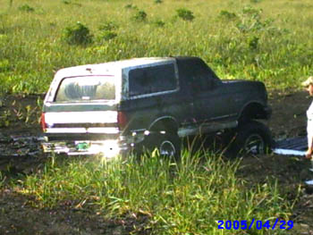 1992 Ford Bronco - Rear three-quarter view of a dark green full-size Bronco with a white top sitting axle-deep in a muddy water hole in a grassy field, its bo…