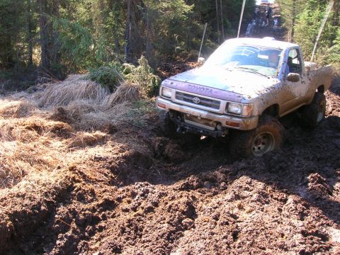 2003 Jeep TJ - A Toyota pickup truck navigates a challenging muddy terrain on a wooded trail, with another vehicle visible further up the path.
