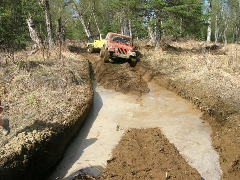 2003 Jeep TJ - Two Jeep Wranglers navigate a challenging muddy trail in a wooded area.