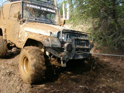2003 Jeep TJ - A heavily mud-covered Jeep Wrangler navigates a rough, wooded trail, with a tow rope attached to the front.