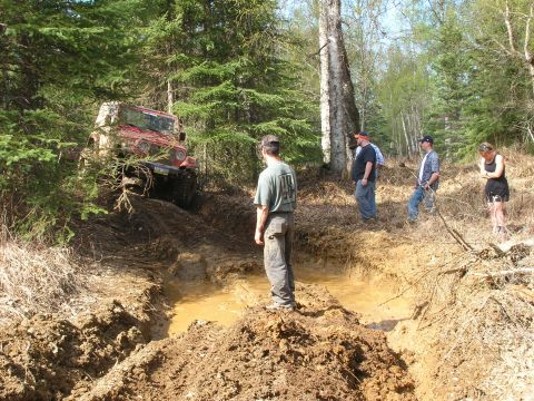 2003 Jeep TJ - A red Jeep Wrangler navigates a challenging muddy off-road course with onlookers observing the action.