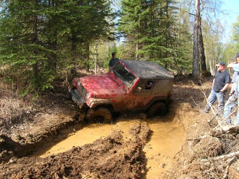2003 Jeep TJ - A red 2003 Jeep TJ Wrangler navigates a difficult muddy obstacle on a wooded trail.