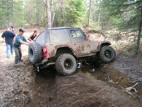 2003 Jeep TJ - A red Jeep Wrangler navigates a muddy off-road trail with assistance from bystanders.