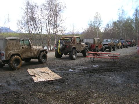 2003 Jeep TJ - A line of muddy off-road vehicles, primarily Jeeps, are parked on a dirt lot next to a body of water.