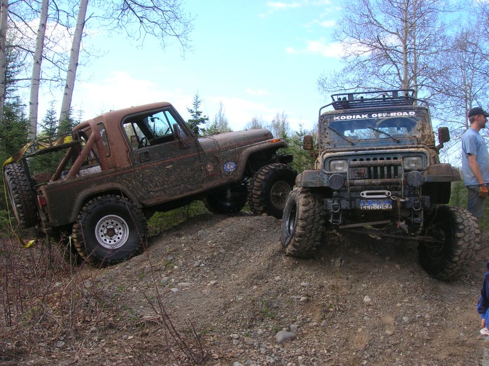 Two muddy Jeeps are parked on a dirt incline. One is a brown CJ-7, the other is a dark green YJ with a roof rack