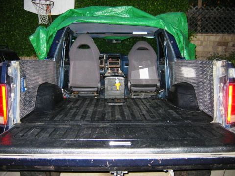 1985 Ford Bronco - The interior of a blue Ford Bronco shows two bucket seats and a covered bed, with a basketball hoop visible in the background.