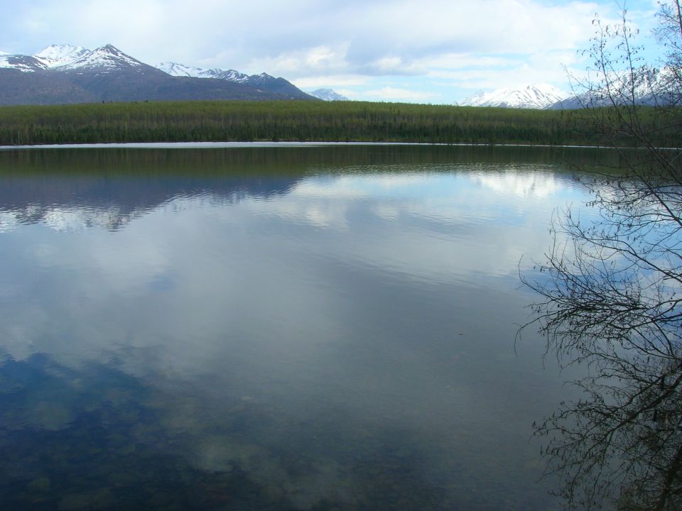 Lake reflects snow-capped mountains and trees under a cloudy sky