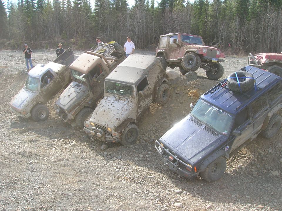 Several muddy off-road vehicles, including a blue Jeep Cherokee and a red Jeep Wrangler, parked on a dirt incline with trees in the background