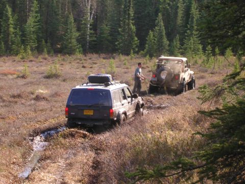 2003 Jeep TJ - Two Jeeps navigate a muddy, wooded terrain, with one vehicle appearing to be stuck in the mire.