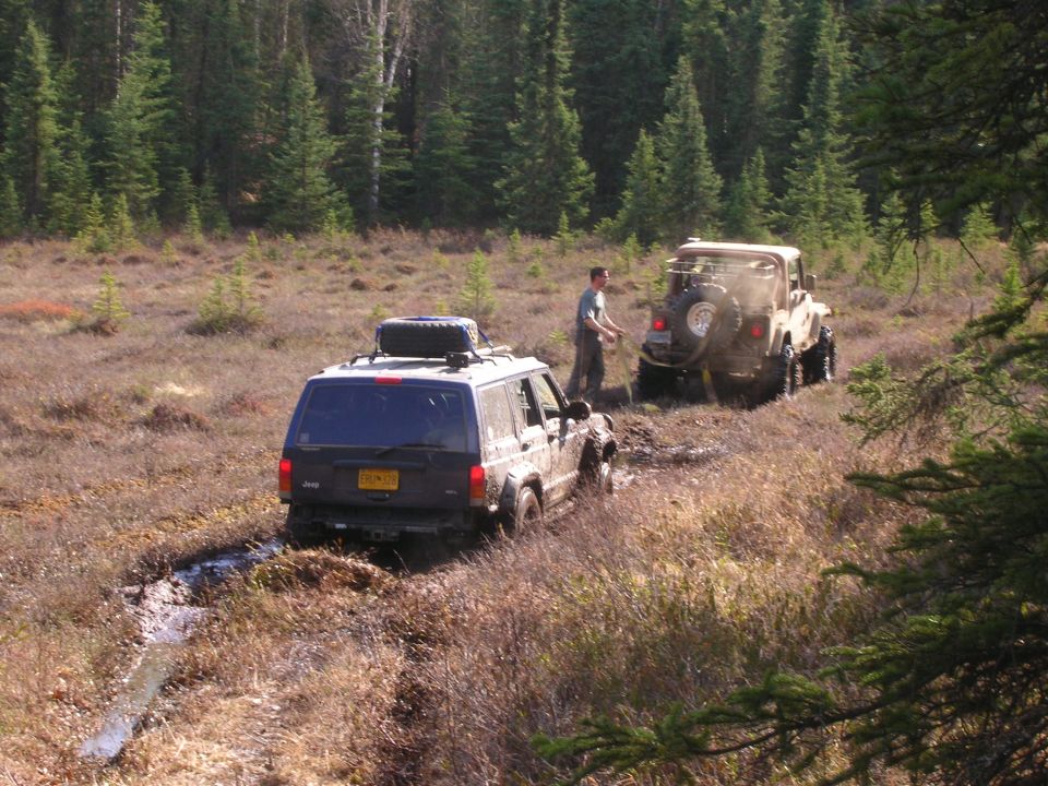 Dark blue Jeep Cherokee is stuck in mud, with a tan Jeep Wrangler behind it. A person is standing between them