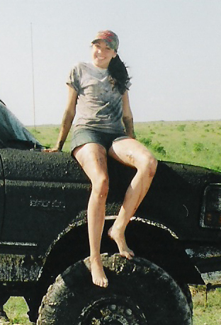 1992 Ford Bronco - A person poses seated on the mud-caked front of a dark off-road SUV, resting bare feet on an oversized muddy tire, in an open grassy field;…