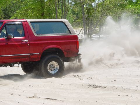 1994 Ford Bronco - A red SUV drives off-road, creating a large plume of dust from the sandy terrain.