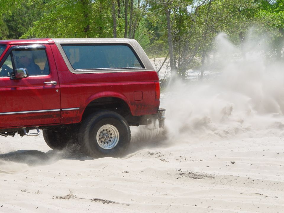 Red SUV with a tan top driving on sand, kicking up a large dust cloud. Visible text: SUPERMOTORS.NET