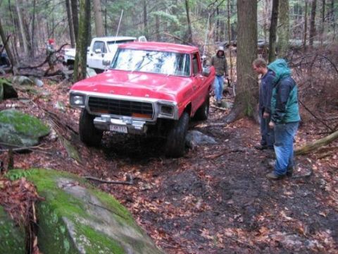 1978 Ford Bronco - A red Ford pickup truck, likely from the late 1970s, is seen off-roading on a muddy forest trail with other vehicles and people present.