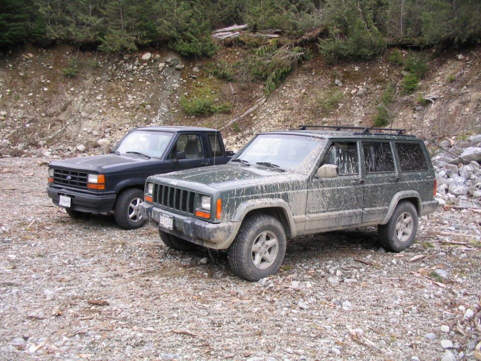 Muddy green Jeep Cherokee XJ and blue Ford SUV parked on rocky gravel below a forested slope