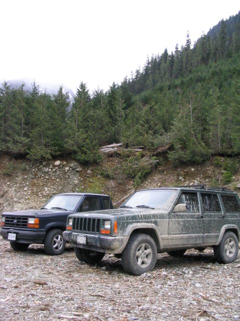 2000 Jeep Cherokee - Two SUVs are parked on a rocky surface with a dense forest and misty mountains behind them.