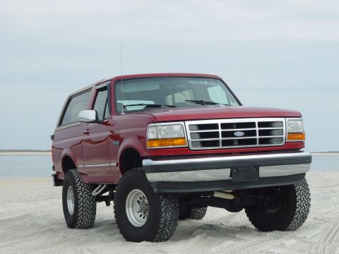 1994 Ford Bronco - This lifted red Ford Bronco is parked on a sandy beach, ready for off-road adventure.