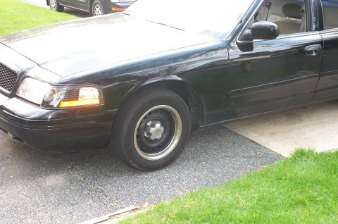 1999 Ford Crown Victoria - Front half of a black Ford Crown Victoria on a driveway, showing the driver-side fender and door with black police-style steel wheels weari…