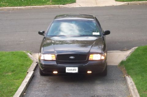 1999 Ford Crown Victoria - Head-on front view of a black Ford Crown Victoria sedan sitting in a residential driveway with headlights and fog lamps lit, showing the bl…