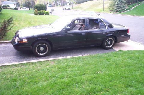 1999 Ford Crown Victoria - Driver's-side profile of a dark green Ford Crown Victoria sedan parked on a suburban concrete driveway, riding on black steel police-style…