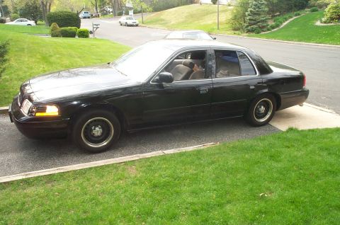 1999 Ford Crown Victoria - Three-quarter front view of a black Ford Crown Victoria sedan sitting low on a concrete driveway in a hilly suburban neighborhood, with tan…