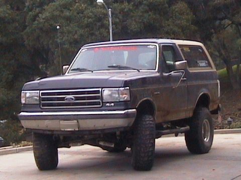 1991 Ford Bronco - This lifted Ford Bronco has a dark blue body and a tan removable top, viewed from the front three-quarter angle.
