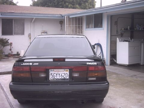 1989 Mazda MX-6 - The rear of a black Mazda MX-6 is parked in a driveway next to an open garage containing laundry appliances.