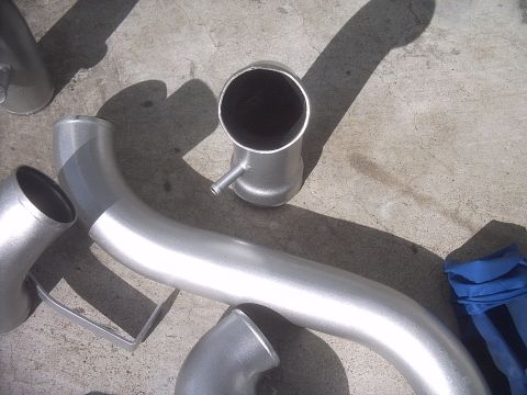 1989 Mazda MX-6 - Several metal exhaust pipes and connectors are laid out on a concrete surface, with a blue cloth visible on the right.
