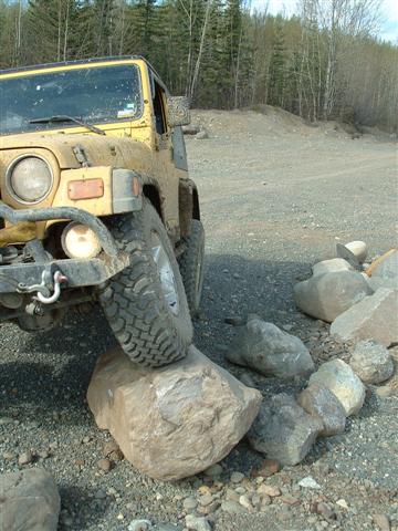 2000 Jeep TJ - A yellow Jeep TJ navigates an obstacle course, with its front tire perched on a large rock.