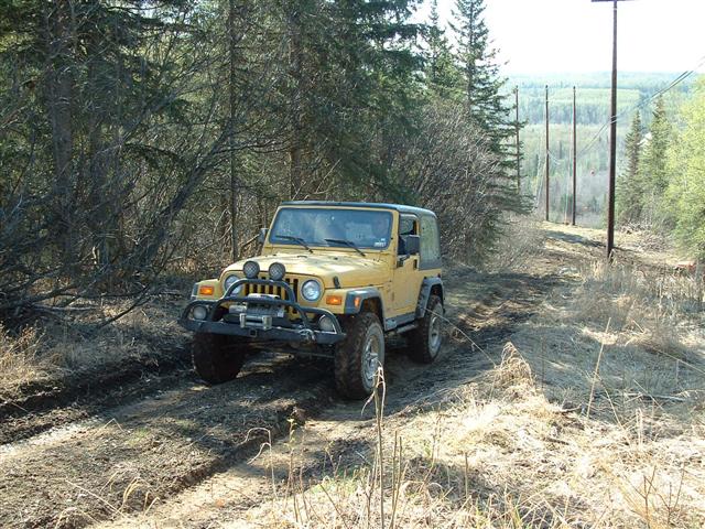 Yellow 2000 Jeep TJ driving on a muddy dirt road through trees