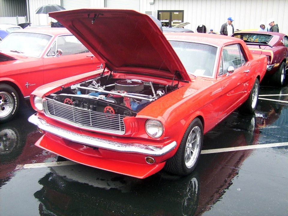 Red 1965 Ford Mustang coupe, hood open, parked on wet asphalt