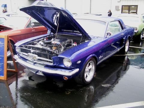 2002 Ford Mustang - A blue 1965 Ford Mustang coupe is displayed with its hood open, parked on a wet surface next to other vehicles.