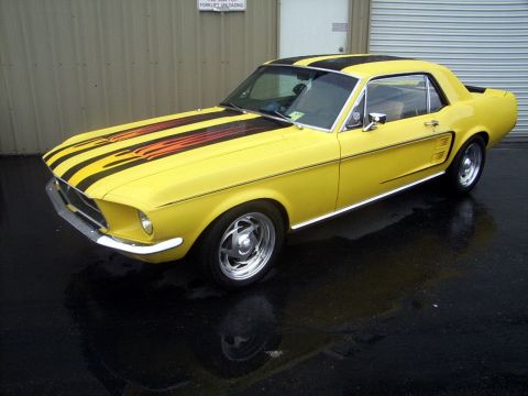 2002 Ford Mustang - A yellow 1967 Ford Mustang coupe with racing stripes is parked outside a garage.