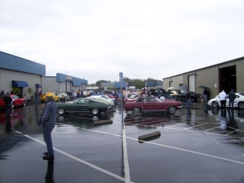2002 Ford Mustang - A collection of classic cars, including a green 1967 Ford Mustang, are parked in a wet lot outside industrial buildings.