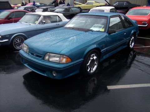 2002 Ford Mustang - A teal coupe with chrome wheels is parked on wet asphalt, surrounded by other vehicles.