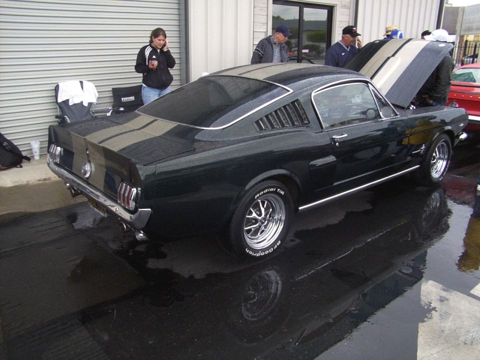 Dark green 1965 Ford Mustang with silver racing stripes, chrome wheels, and louvers on the rear quarter windows