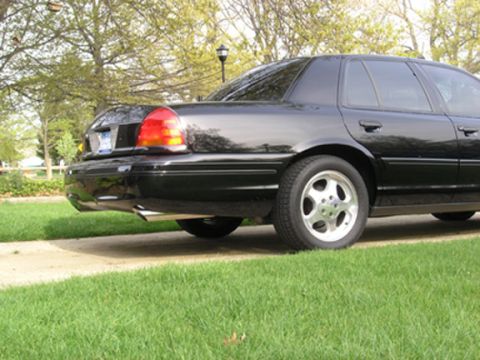 1999 Ford Crown Victoria - The rear of a black sedan is shown parked on a lawn with trees in the background.