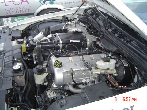 1999 Ford Crown Victoria - Close-up of a V8 engine bay a supercharger and other engine components. Another vehicle is partially visible.